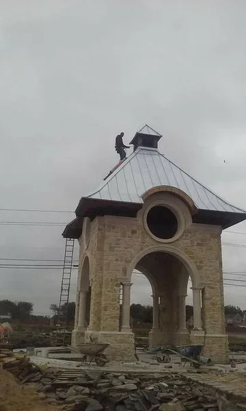 Standing seam metal roof installation on stone tower with peaked pyramid design and circular windows