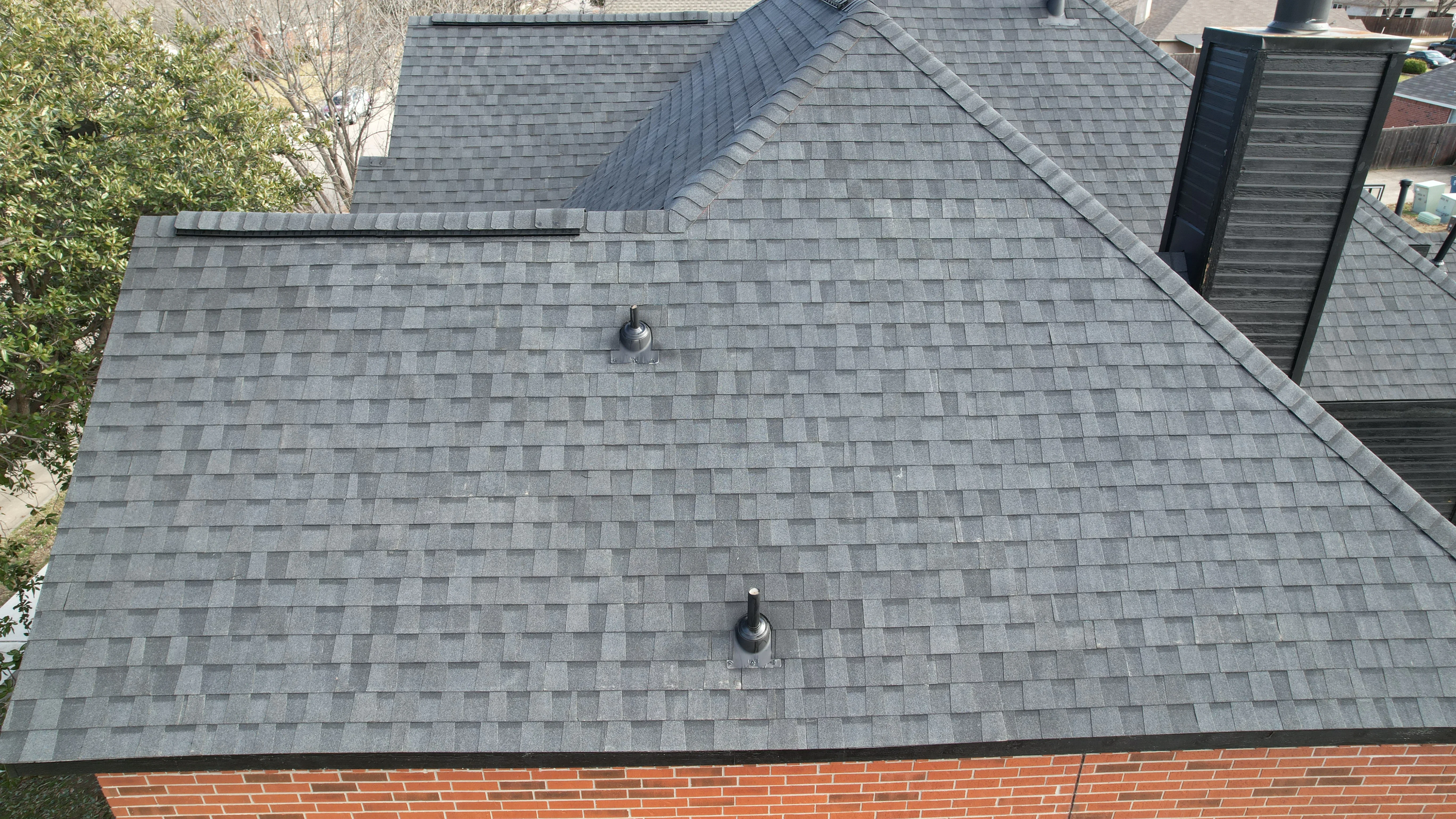 Slate gray architectural asphalt shingle roof with ridge caps and plumbing vents on brick home