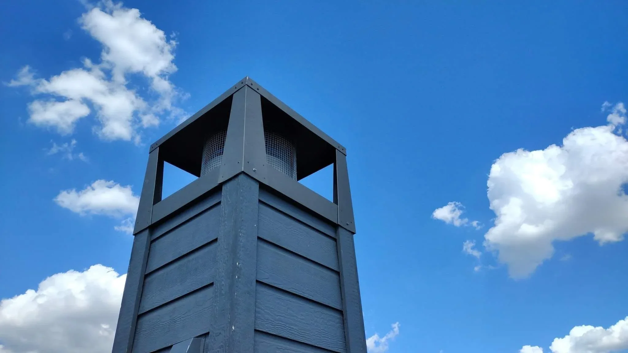 Tall metal tower with peaked roof, ventilation louvers, and horizontal metal siding panels against blue sky